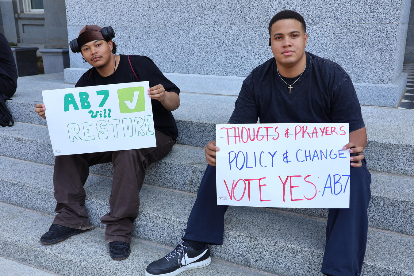 Two young advocates sit on stone steps holding signs reading Vote Yes on AB7.
