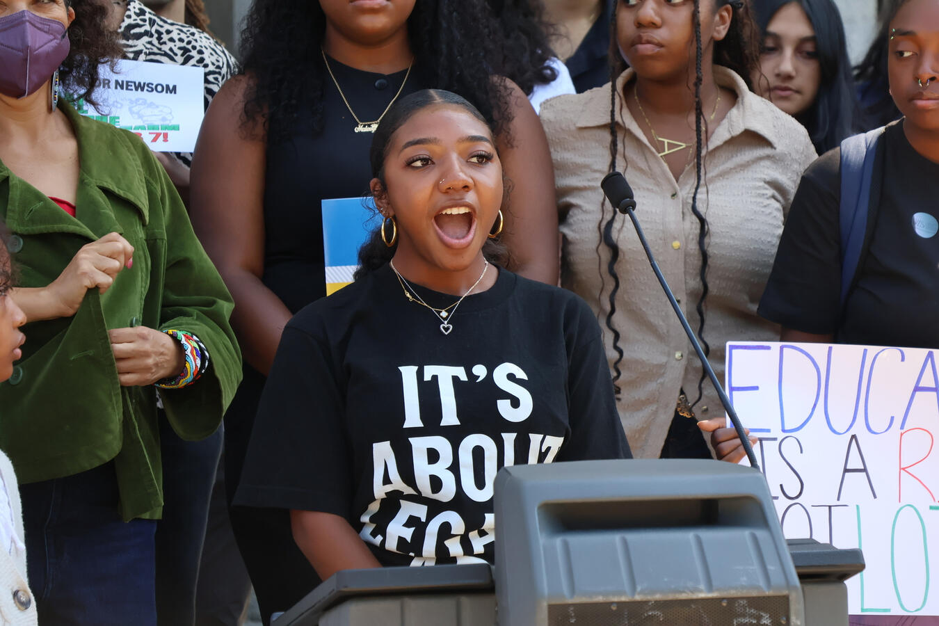 Advocate Speaking in Support of AB7 A woman in a Legacy campaign shirt speaks passionately at an indoor lectern.