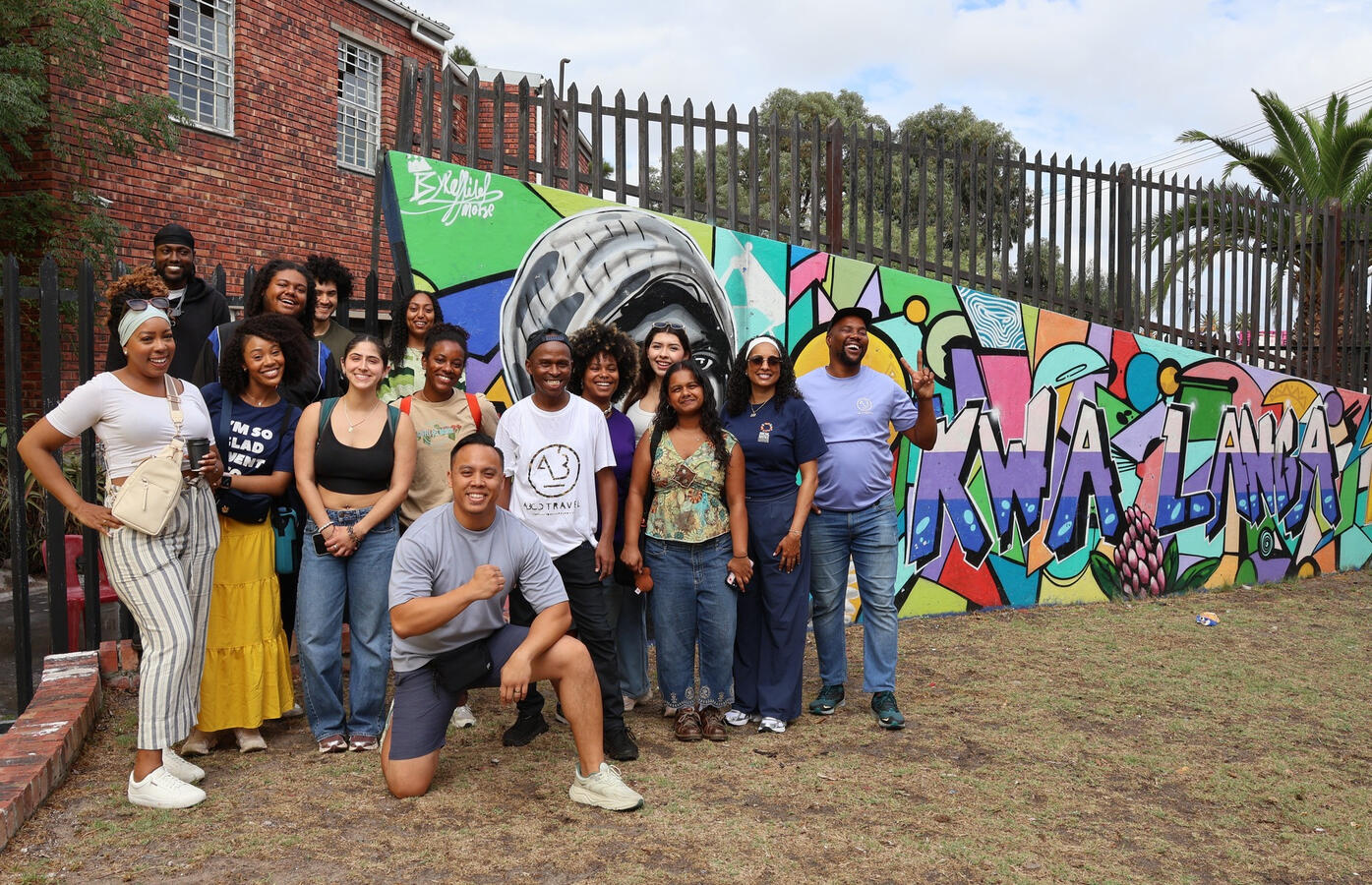 The delegation poses together in front of a large colorful mural on a brick wall.